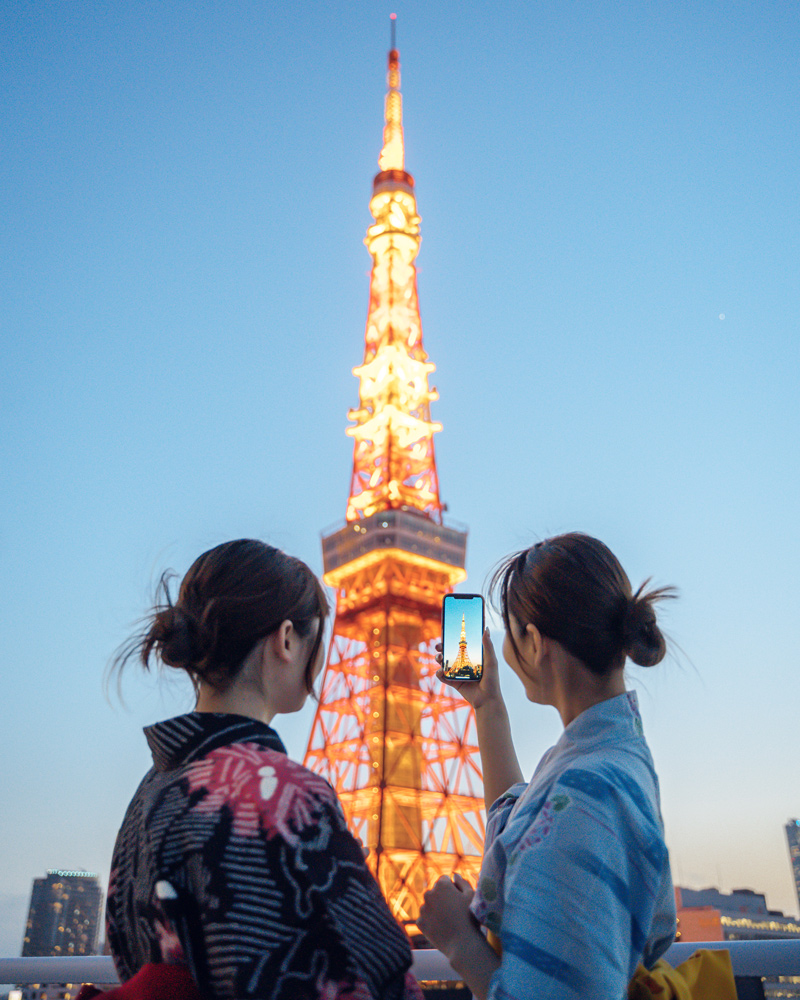 Two women in kimono viewing an illuminated tower at dusk from a rooftop