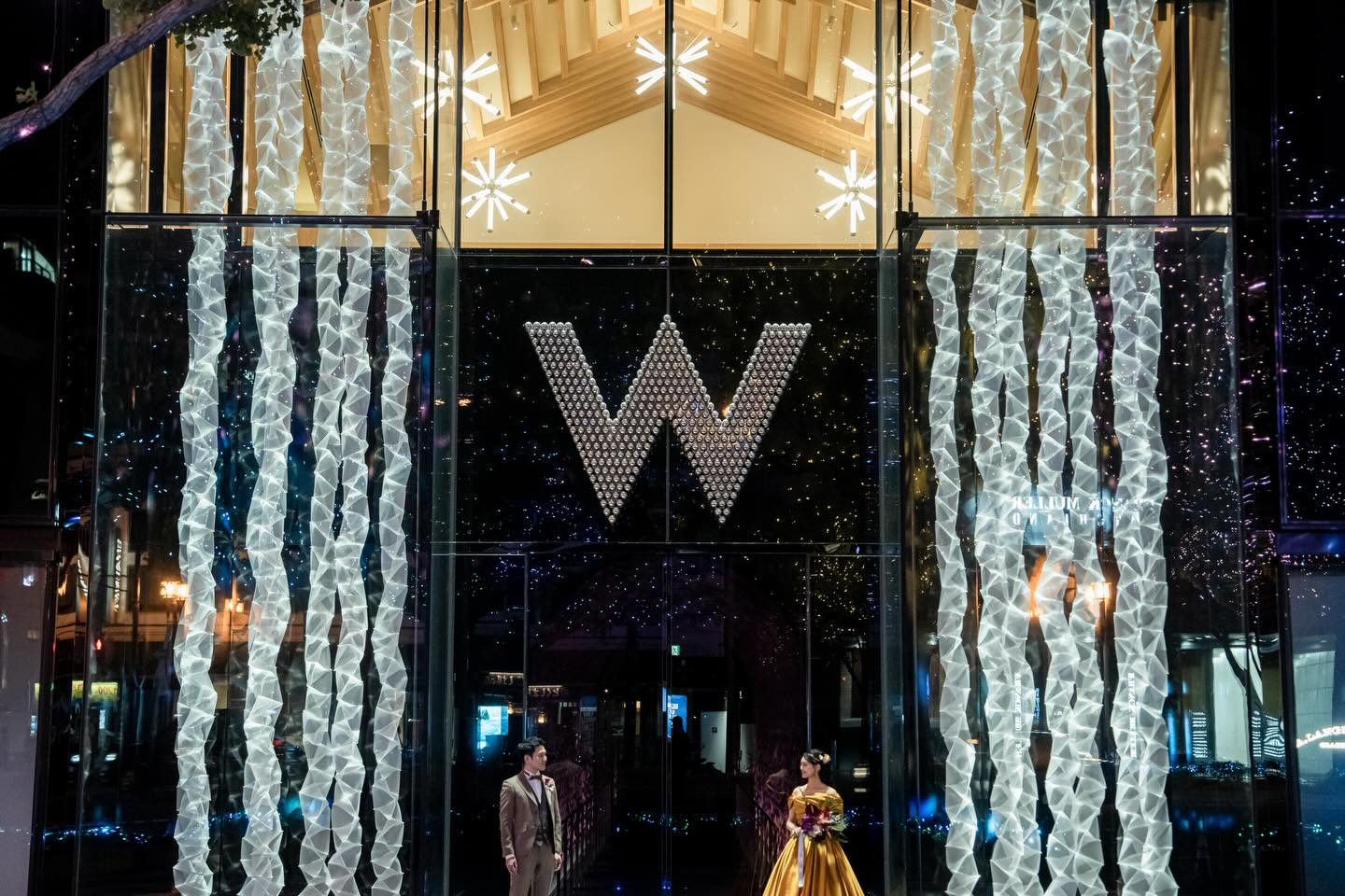 W Osaka's illuminated glass entrance at night, with a couple in formal wear posing beneath the lit 'W' sign